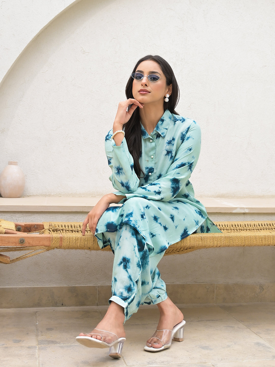 Woman wearing a light blue floral print outfit and sunglasses, seated on a bench.